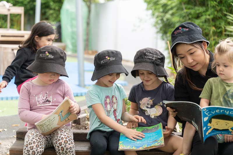 Four children sit outdoors holding books and having fun learning with an educator. One child sits on the educator's knee reading where they share a book. Next to them, two children share a book. Another child to their left reading another book. Books and interests can bring children together to make new connections and discover childhood friendships.