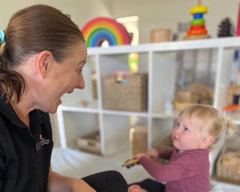 Educational Leader sits cross legged in front of a young child with shelving in the background filled with colourful and natural resources..