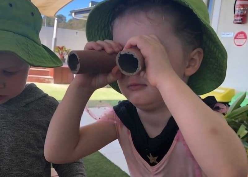 A child in a pink and black shirt and green hat plays in an outdoor environment with a pair of scrap circular tubes a household recycling item that she uses for pretend binoculars.