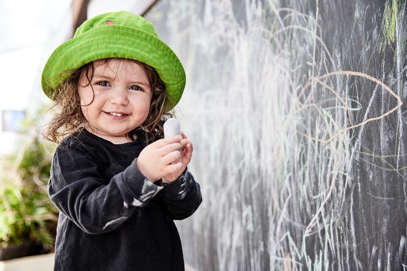 A toddler in a green hat holds a big fat piece of homemade chalk, a perfect ideal stocking stuffers idea. The child stands in front of a blackboard which is covered in chalk scribbles.