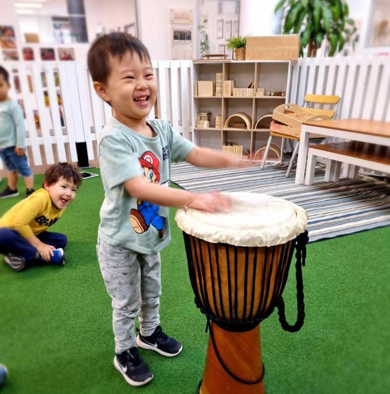 A happy child stands behind a large drum using the beats for storytelling through song and music while behind him two children listen and watch.