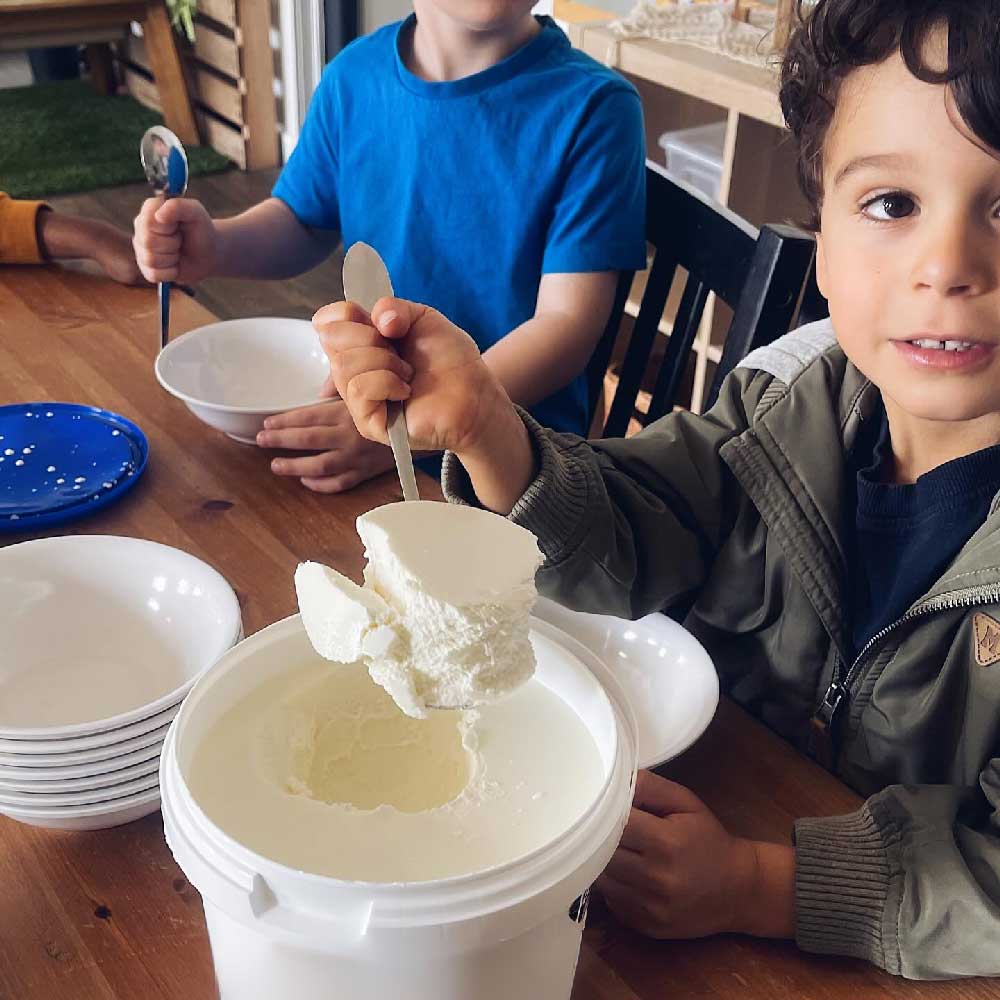 Children sit together at a table, spooning yohurt into their bowls. The child nearest the camera holds up a big spoon filled with yoghurt.