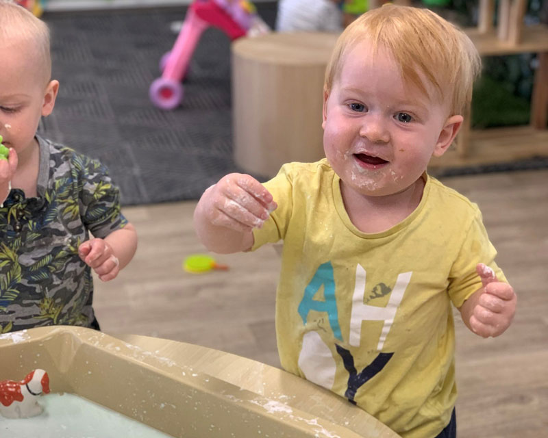 A child in a yellow shirt stands at a sensory table filled with goop. Another child stands to his left playing with the loose parts in the goop.