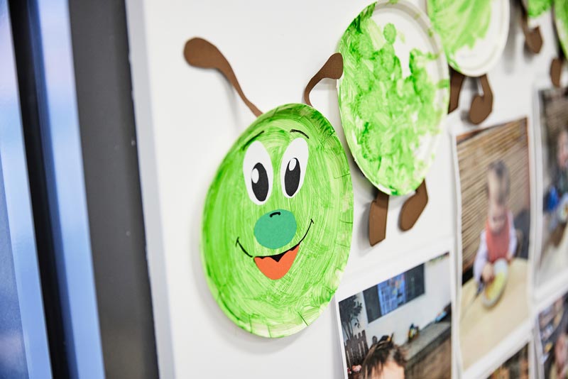 Green-painted paper plates laid out on a wall to form the hungry caterpillar. The first plate looks like a mask with the caterpillar's face. It has big googly eyes, a darker green nose, a smile with a red tongue and brown antennae. Other green plates have brown feet. Beneath are photos of children eating.