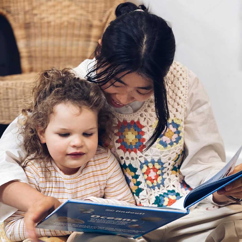 An educator sits on the ground with a child. They are connecting over a book titled, Oceans. The educator is reading to the child while the child points at the images and words. Behind them is a cane chair.