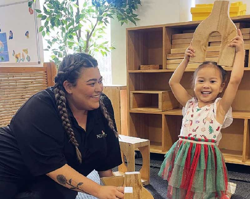 Educator connects with child in indoor play area. the child wearing a red and green tutu-like skirt is holding a y-shaped wooden block above her head. She is grinning. The educator is sitting on the floor at the child's level and has one hand on a wooden block on the ground. Behind them is wooden shelving with more wooden blocks.