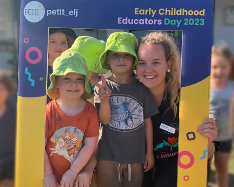 Lead Educator, Samantha poses with children inside a cut-out corflute sign with Petit ELJ and social media branding showing how much fun she is having with the children on early childhood educators day.