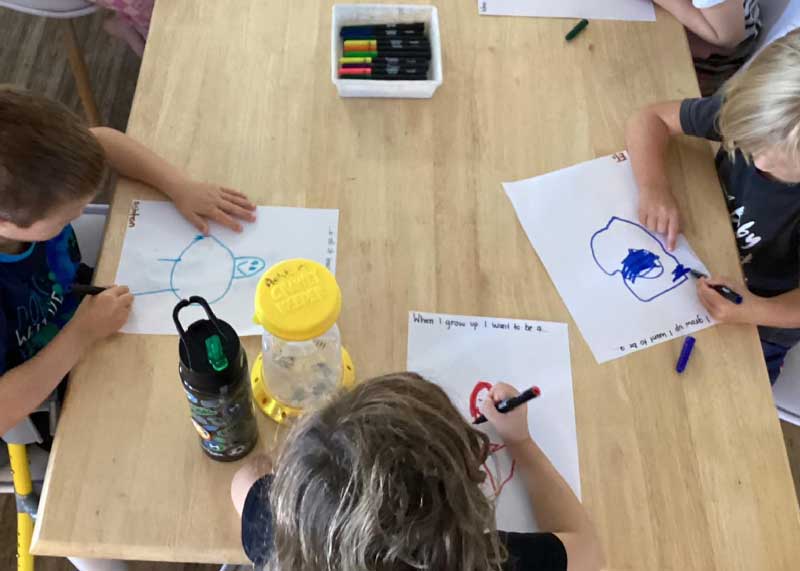 Three children sit a table in a childcare centre, exploring their identity. They each have a piece of paper with the question, "When I grow up, I want to be a...". The children hold a coloured marker to draw their answer. The child in the middle has a bug container next to him and a black waterbottle. Encouraging children to take water with them can help reduce dehydration.