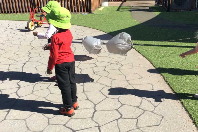 Two children play with "home-made" kites made from plastic bags on pavement in an outdoors setting. There are four shadows of children on the ground and a tricycle in the background near a wooden fence.