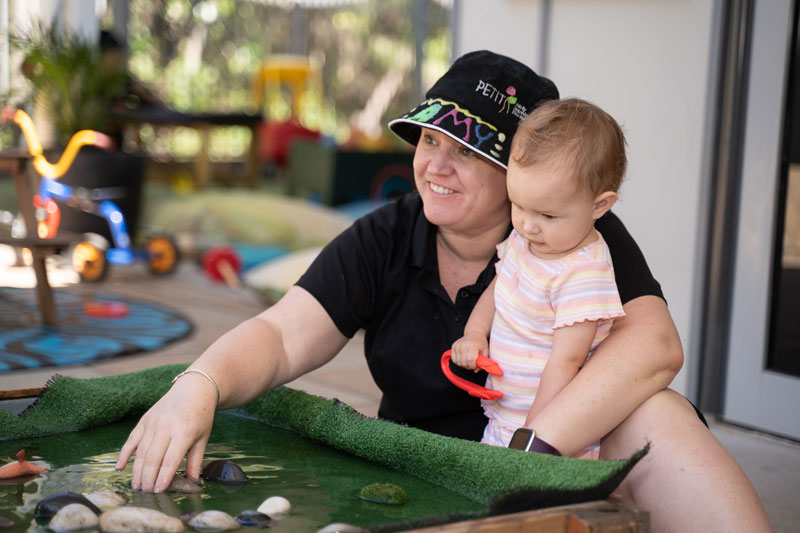 An educator sits in a shaded outdoor setting, dipping her hand in a table lined with fake green grass, over which water flows around pebbles for a sensory water experience. A child stands in the educator's arms watching and waiting to enjoy the experience.