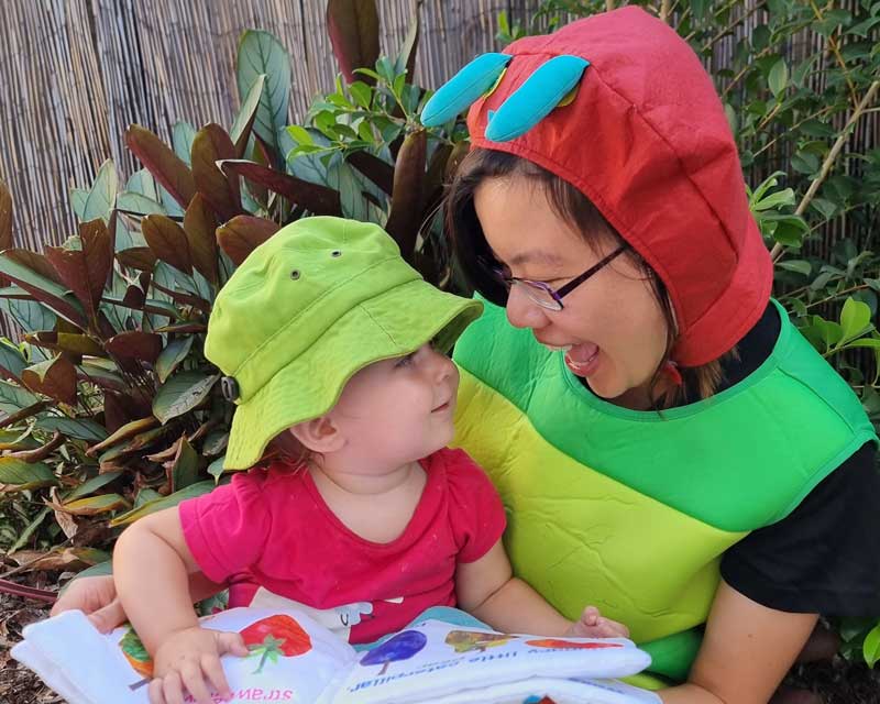 A child holds a picture book about The Very Hungry Caterpillar. They sit on the lap of Educational Leader Bao Huynh who is dressed up like the book's main character.