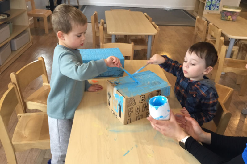 Two children, one standing, one sitting at a table work together to paint a memory box from repurposed cardboard, blue.