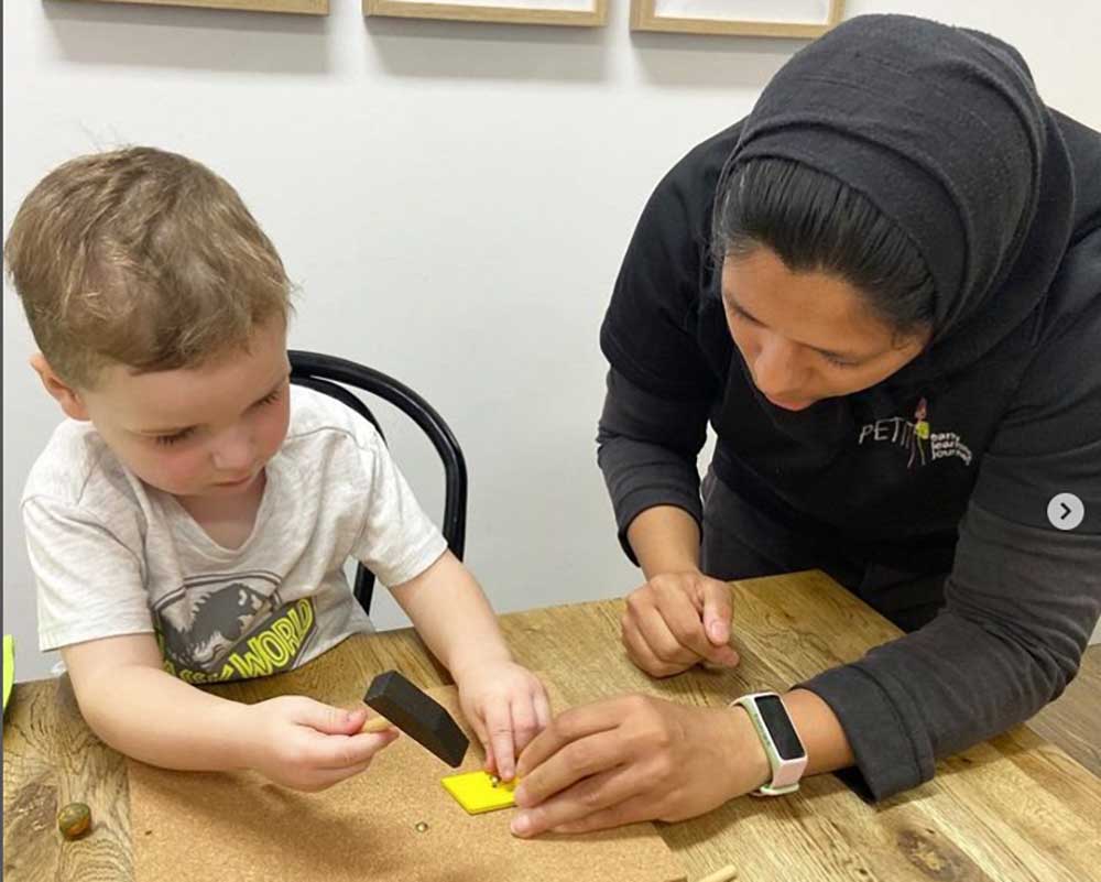 A child sits at a desk participating in a studio inquiry project. On the table in front of him is a cork board with tacts. He holds a tac over a yellow rectangular shape. In his right hand is a small hammer. An educator leands in to the table and holds the board for the child.