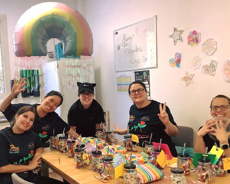 Petit ELJ Wooloowin team smile and laugh at the camera as they gather around a table with jars and colourful stripped packages as they celebrate Educators Day. The educators are wearing customised early childhood educators day shirts. Petit's People and Culture Operations Manager makes the peace sign as she contributes to employee engagement.