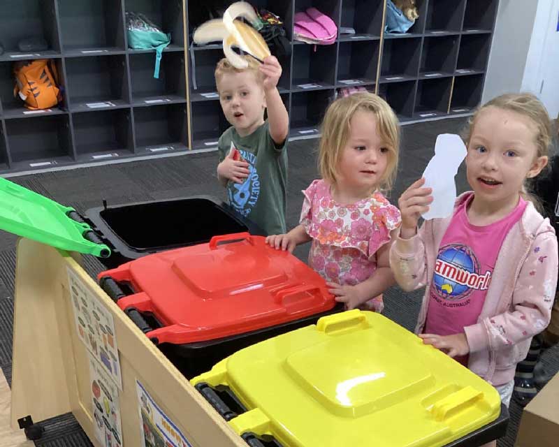  Three preschoolers inside a classroom stand in front of colourful recycle bins: green, red and yellow. They are playing an indoor game and learning a children's chore activity for home, sorting out the rubbish into the correct bins.