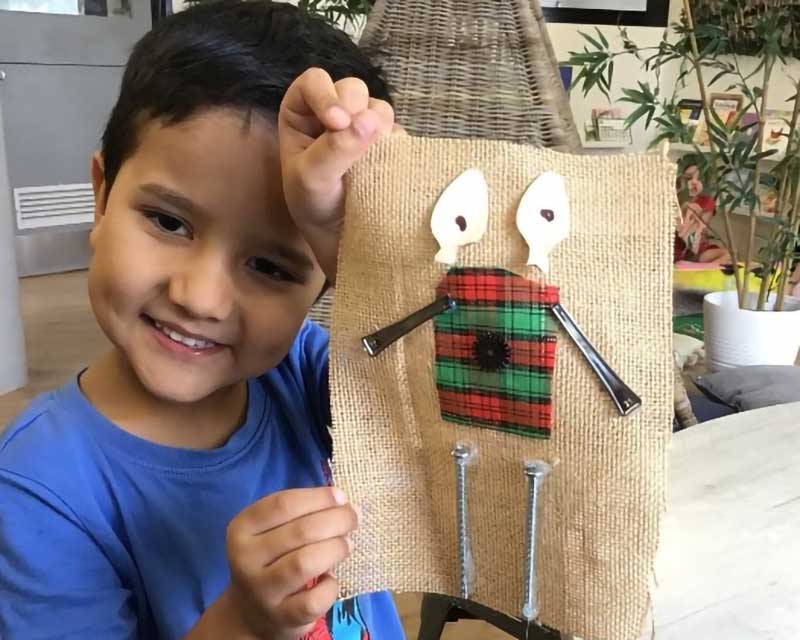 A child holds up an art piece with an image that represents his identity made with loose parts and recycled things such as long screws, pieces of material and a button. He tells stories through art.
