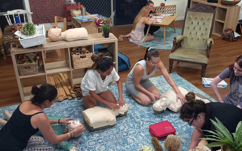 A group of educators kneel over dummies to practise their CPR skills to maintain their annual qualifications as part of their professional development planning at a training evening at Petit ELJ Caloundra. In the background two educators complete first aid documentation.