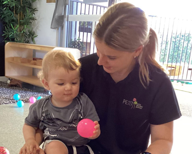 An educator sits with a child in a shaded environment, giving them the room to express their feelings. The child sits on the educator's knee and holds a pink ball. The educator is listening and encouraging the child to talk about their feelings. In the background is a entranceway to an outdoor playing area. Coloured balls are scattered behind them.