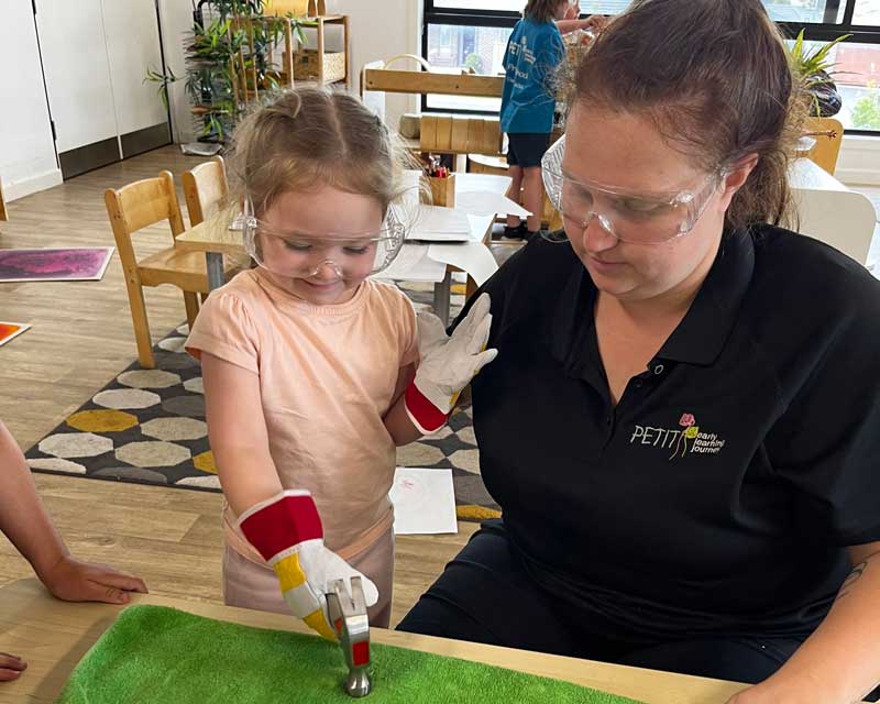 Shannon Ivanovski wears protective glasses as she helps a child wearing another pair of protective glasses who stands next to her with a hammer as they work together on a art project.