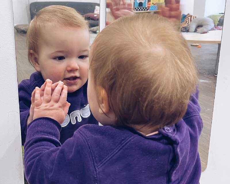 A baby plays with a wall mirror activity, standing up with one hand flat and touching the mirror and the other waving. The child is reflected back and in the background are comfy spaces with cushions and chairs.