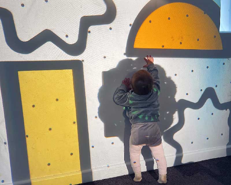 A child reaches up with a hand where shapes are cast against a wall. Manipulating shapes in light and shadow games helps to improve fine motor skills.