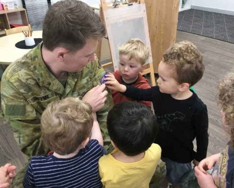 A dad wearing his Australian army uniform crouches down while visiting his child's centre. He is surrounded by five children who take an interest in the Australian flag sewn onto his shirt. He shares information about his role and the army through his parent involvement.