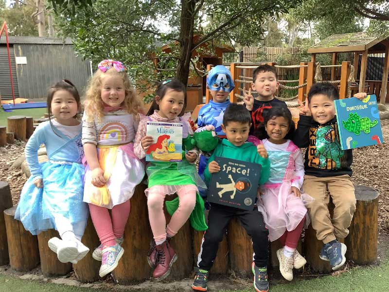 A group of children pose under the shade of a tree for CBCA Book Week in their favourite character costumes. Three children also hold the books that inspired their imaginations at Petit Early Learning Journey.