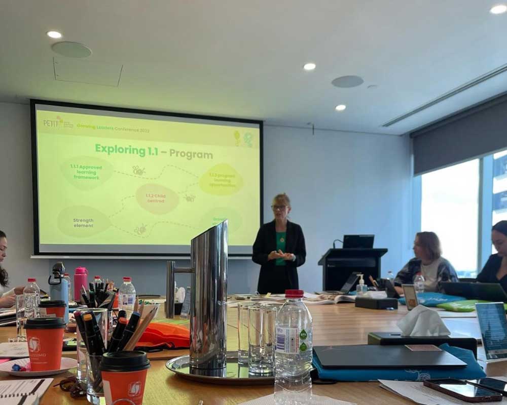 A projector screen in the background displays a powerpoint slide with the words Exploring 1.1 - Program under the title, Growing Leaders Conference 2023. A presentor stands next to the screen and beside a lecturn. The table in the foreground has many different items spread across it include coffee cups, water flasks, bottles of water, pens, pencils and markers. To the right and left of the image, educators look engaged in the prsentation which will cover inquiry-based learning.