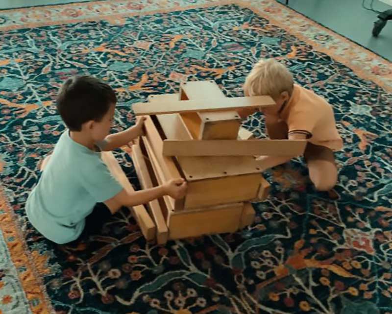 Two preschool children participating in an indoor activity, crouch down low on a large mat playing. They are playing with an assortment of large wooden blocks. A child in a green shirt is holding a wide wooden block up and on the other side, a child in a pale orange shirt looks closely at their construction.