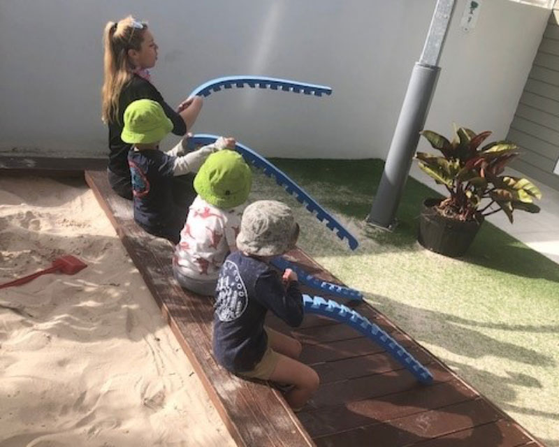 An educator joins in the role-play of going fishing. She holds a pretend foam fishing rod and next to her are three other children who are all fishing too. There are many different types of imaginative play including role play and pretend play.