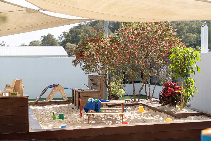 A Petit Early Learning Journey sandpit play space basks in filtered sunshine beneath a canvas sail. One the sandpit are several colourful buckets and spades. In the centre is a wooden table and to one side a wooden sand play kitchen. The right side of the sandpit has a few shrubs and trees.