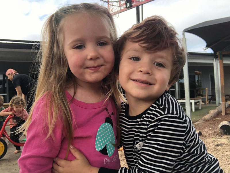 A child in a pink long-sleeved shirt and another child in a white and black horizontal long-sleeved shirt hug in an outdoor play environment as they learn to build social skills and find friends. Behind them, a child is hopping on a red tricycle with black and yellow wheels.