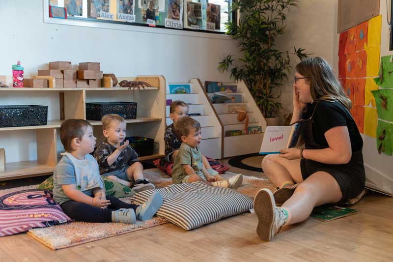 An educator reads and learns about herself as she engages a group of toddlers who are sitting on cushions as they listen to her storytelling. 