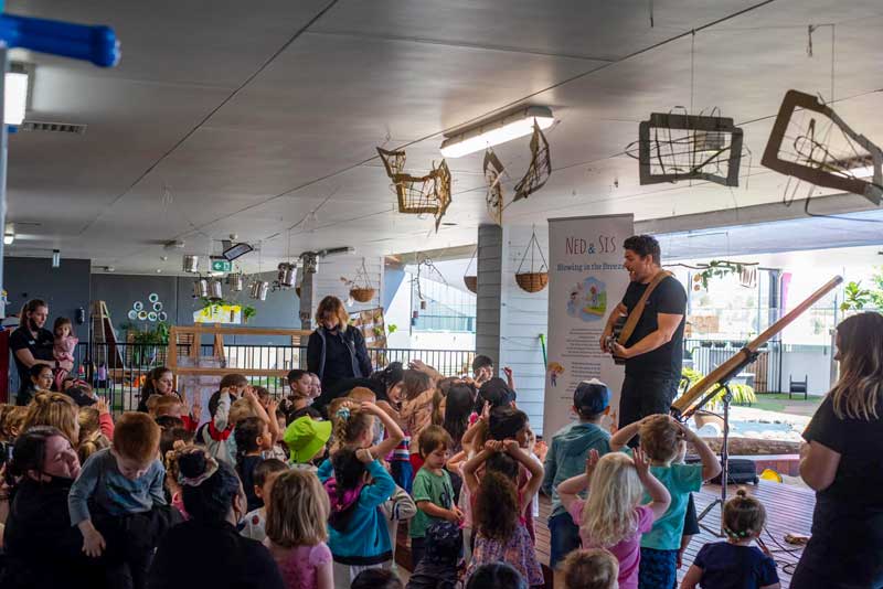 Many children gather before a performer on the stage. The children's performer holds a guitar and he is singing to his audience who share his storytelling through movement and dance by placing their hands on their heads.