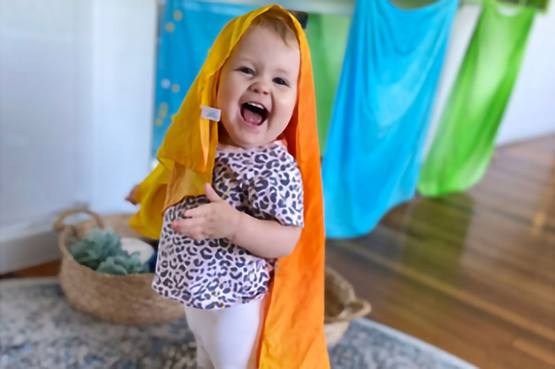 Toddler laughs facing camera with bright orange material - possibly light enough to make a children's kite fly - covering their head and back.