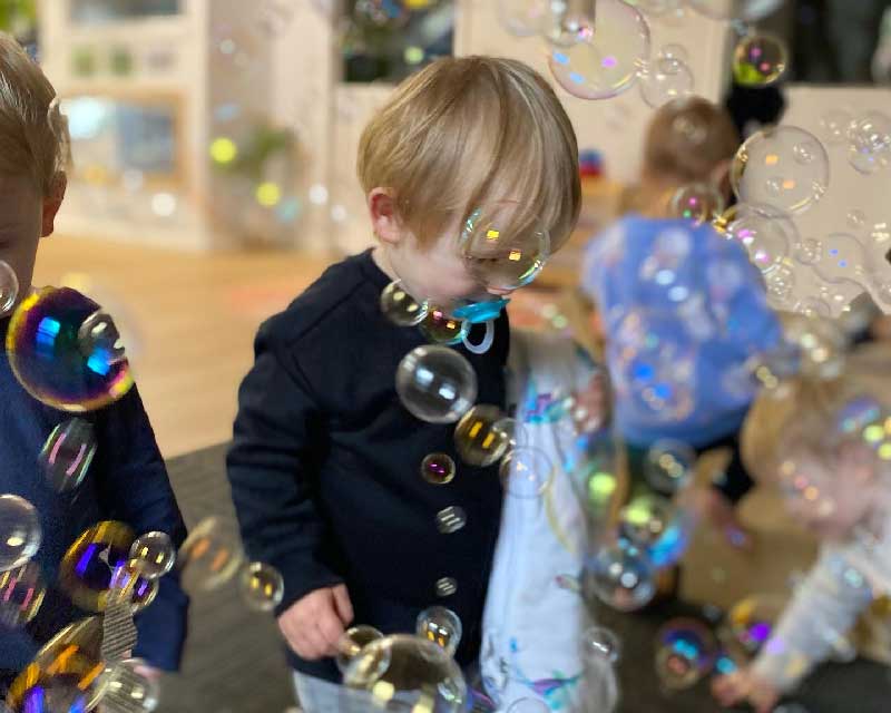 Group of babies and toddlers playing an indoor physical game on a mat surrounded by lots of colourful bubbles. Centre focus on one child holding a blanket with blue dummy in mouth