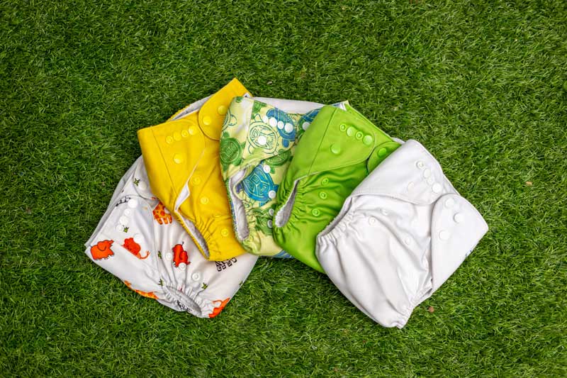5 Colourful cloth nappies displayed in a half-circle in front of a synthetic grass background.
