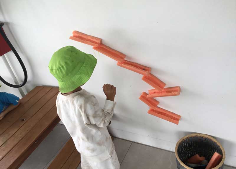 A recycled marble run! A child dressed in a white tracksuit watches a marble fall through a wall-installed recycled marble run made with tubular soft plastics.