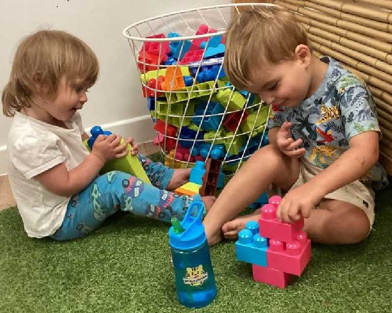 Two children sit together in the corner of a daycare centre playing with big plastic blocks. They sit on artificial lawn and behind them is a basket full of blocks. Between them is a blue water bottle with a green straw, clearly a favourite accessory that encourages a child to drink more water. 