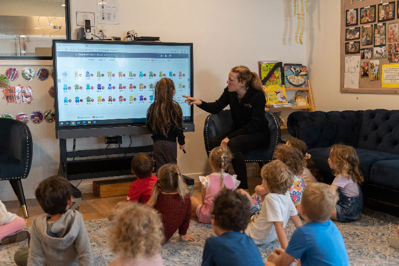 An early childhood educator sits in front of an electric white board inside a studio. The white board has multiple colourful emojis representing emotions. A child stands in front of the board holding a pen while the Petit ELJ educator encourages a group of preschoolers to recognise their emotions. Introducing an educational program and practice that supports children's health and wellbeing is an important aspect of the NQF. 