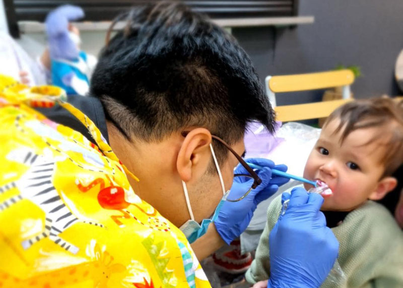 Dentist visiting an early childhood centre peers into a young child's mouth. The dentist holds a mirror and light. The child wearing a green shirt is calm and looking towards the camera while enjoying their first dental visit.