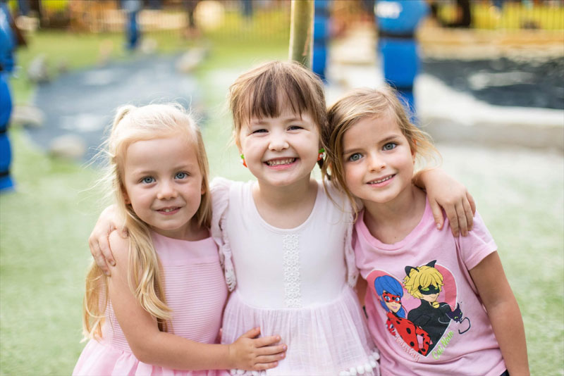 Three children happy and smiling at the camera. Behind them is an outdoor environment with many engaging experiences to help support their wellbeing and cope with change.