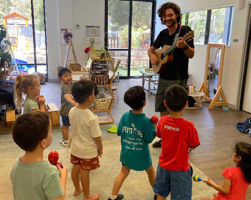 Preschool children hold maracas while singing along with favourite nursery songs. A musician holding a guitar stands facing the children and playing melodies.