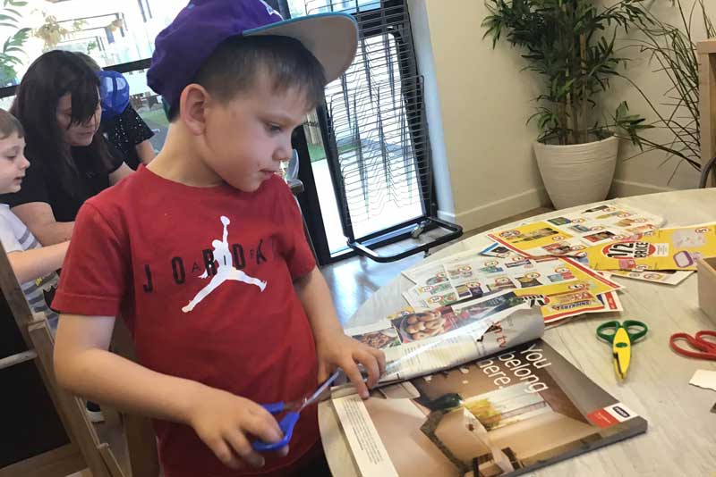 Child in a red shirt stands at a table looking at a glossy magazine. He holds scissors in his right hand preparing to cut the paper to use in making a kite our of repurposed materials.