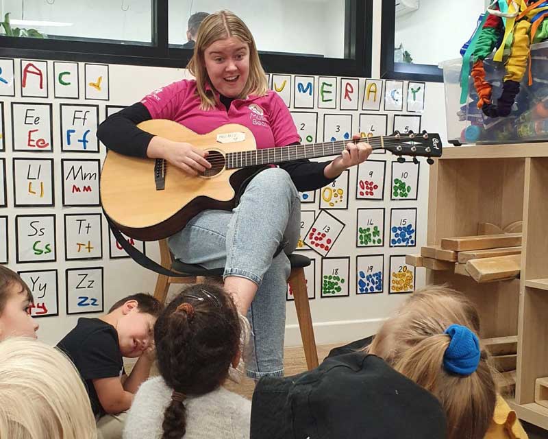 Music beat teacher sits and plays the guitar singing songs with a group of children sitting on the floor. and enjoying their extracurricular experience.