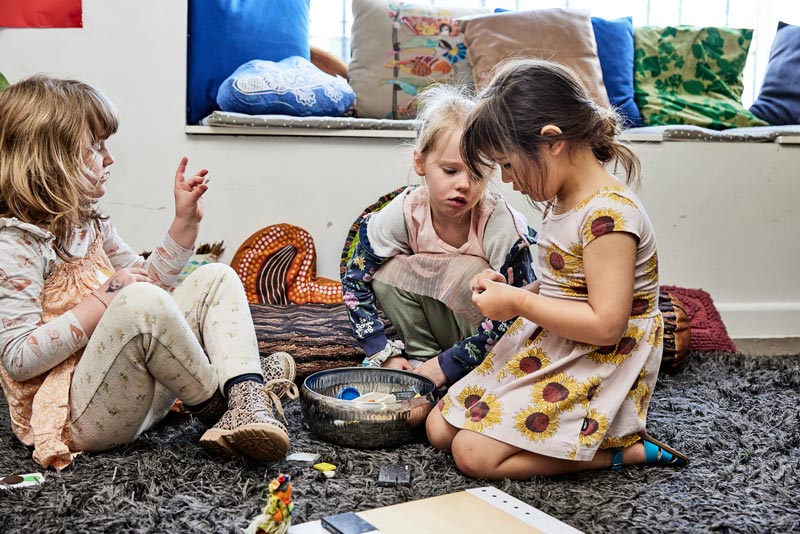 Three children sit on the ground in a circle around a basket filled with loose parts such as lid tops. Lid tops are useful for homemade games, fun stocking stuffers.