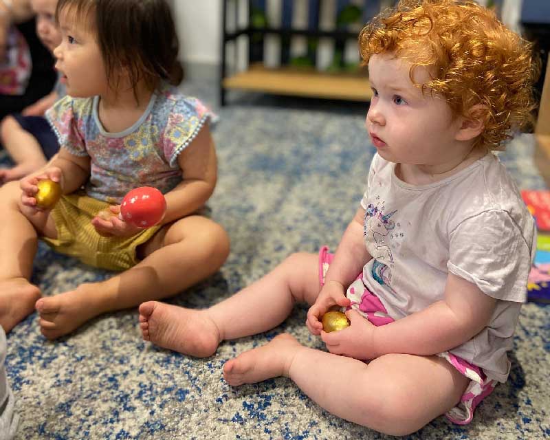 Two children sit a circular carpet listening and singing to children's rhyming songs. One child holds a shaker in their hand.