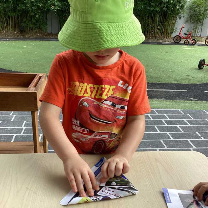 A child in an orange printed shirt and green hat stands at a table and reuses scrap paper for an origami experience. Recycling at home is important as it engages children's curiosity about the world and how to create a sustainable future.