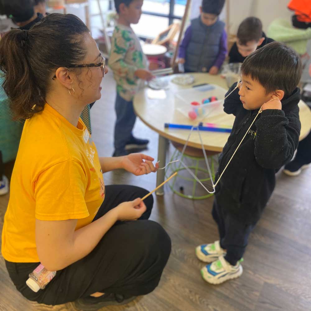 A guest from a science incursion supports children's studio-based inquiry learning activities. Shesits on the floor in front of a child. She holds a metal coathanger. A child stands in front of her. In both hands he holds a piece of string up to each of his ear. The strings are attached to either side of the coathanger. 