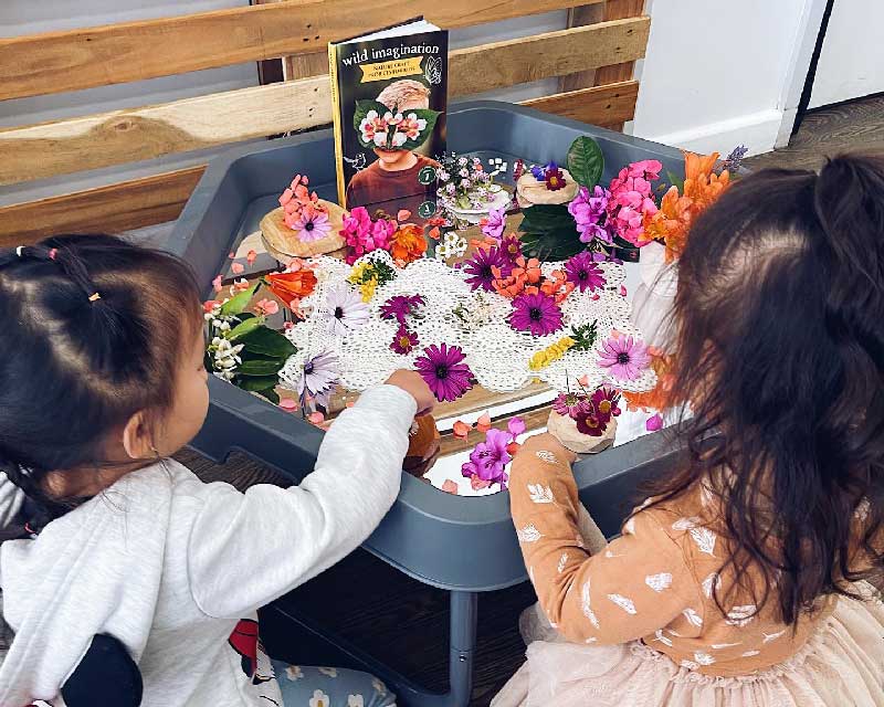 Two children stand in front of a sensory table filled wtih recycled flowers, doyles, pot pouri, wooden things and a book that stands up towards the back. The book is titled, Wild Imagination. It features an image of a child wearing a mask made out of leaves and flowers.